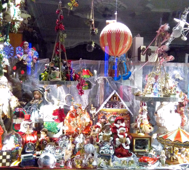 Ein Schaufenster voller Weihnachtsdekorationen, darunter Puppen, Spielzeuge, ein Haus, eine Karussell und andere festliche Gegenstände.