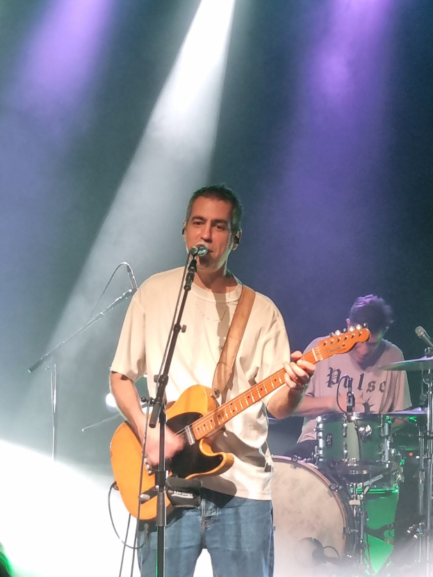 Ein Mann spielt eine E-Gitarre auf der Bühne und singt in ein Mikrofon, während ein anderer Musiker im Hintergrund Schlagzeug spielt.