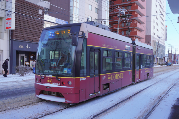 Ein roter und gelber Zug fährt durch eine verschneite Straße mit hohen Gebäuden, Strommästen, Schildern und einer Gruppe von Menschen unter einem bewölktem Himmel.