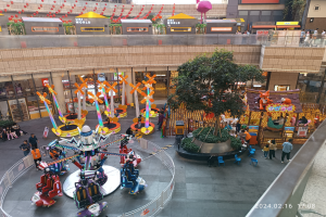 Ein Vergnügungspark innerhalb eines Einkaufszentrums mit verschiedenen Fahrgeschäften, Bäumen, Menschen und Ständen, umgeben von Gebäuden oben und einem Wasserzeichen unten in der rechten Ecke.