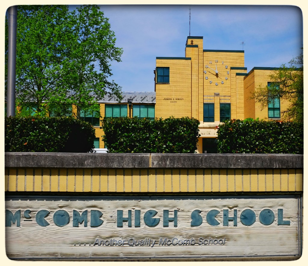 Ein Plakat mit dem Text "MCCOMB HIGH SCHOOL" auf einer Wand, mit Pflanzen, Bäumen, einem Pfahl, einem Gebäude und einem bewölkten Himmel im Hintergrund.