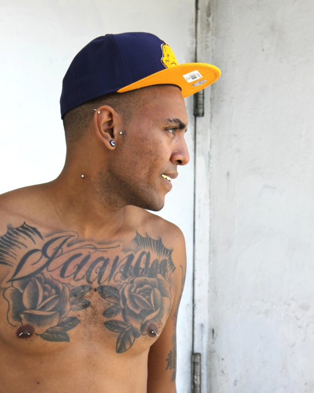Eine Person mit einem Hut mit einem Aufkleber mit einem Strichcode und Bildern, mit sichtbaren Tattoos auf dem Körper, vor einer Tür und einer Wand stehend.