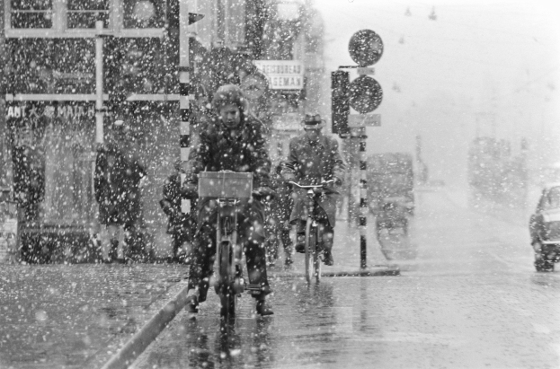 Schwarzes und weißes Foto von Menschen, die auf Fahrrädern eine schneebedeckte Straße entlangfahren, die von Gebäuden, Pfählen und Ampeln gesäumt ist.