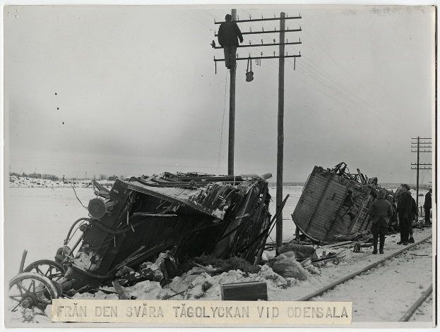 Ein Schwarz-Weiß-Foto eines Zugunglücks auf den Schienen mit ein paar Menschen, die rechts daneben stehen, Pfählen, Drähten, Bäumen und dem Himmel im Hintergrund und Text unten.