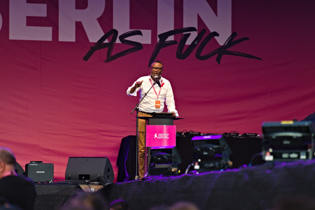 Ein Mann in einem weißen Hemd und braunen Hosen steht auf einer Bühne und hält ein Mikrofon in der Hand, während er vor einem Publikum spricht. Im Hintergrund ist ein pinkfarbenes Banner mit der Aufschrift "Berlin as Fuck" zu sehen.