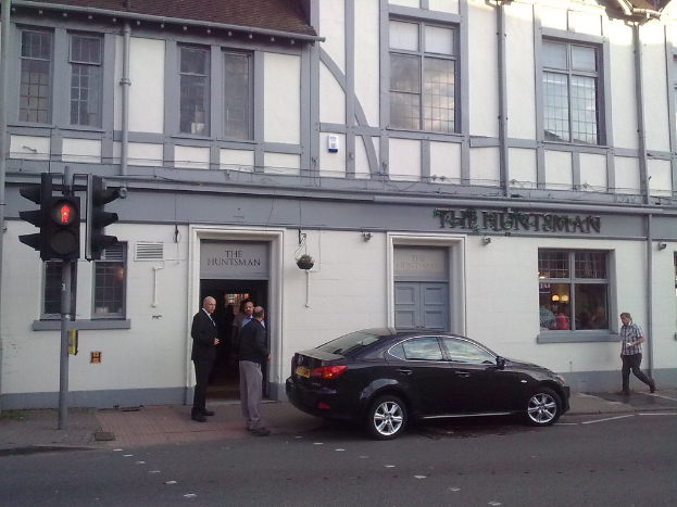 Ein Gebäude mit Text und Rohren an der Wand, ein Auto auf der Straße im Vordergrund, drei stehende Personen, ein Mann auf dem Gehweg, eine Gruppe von Menschen hinter einem Fenster und ein Verkehrspfahl auf der linken Seite des Gehwegs.