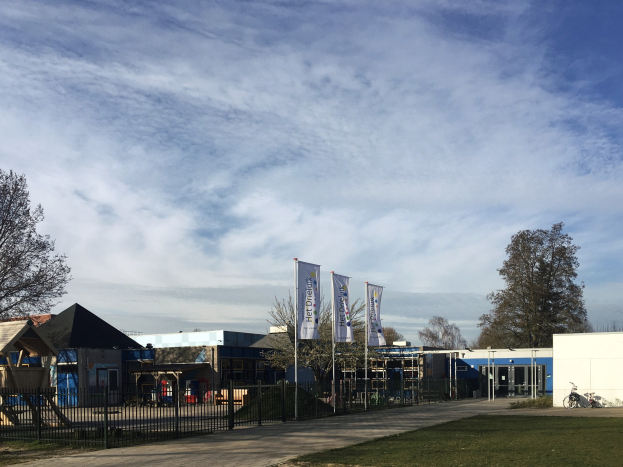 Gro├če Geb├Ąude mit einem grasigen Bereich davor, umgeben von einem Metallzaun, mit einem Weg, B├Ąumen, Bannern, einem geparkten Fahrrad, n├Ąheren H├Ąusern und einem bew├Âlktem Himmel, als das neue Kinderspielgel├Ąnde an der Schule identifiziert.