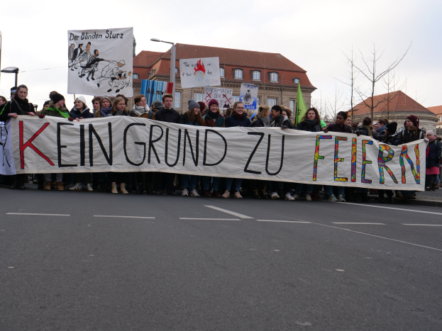 Eine Gruppe von Menschen steht auf einer Straße und hält ein Schild mit der Aufschrift "Kein Grund zu Feiern" hoch, im Hintergrund sind Straßenlaternen, Bäume, Gebäude mit Fenstern und ein klarer blauer Himmel zu sehen.