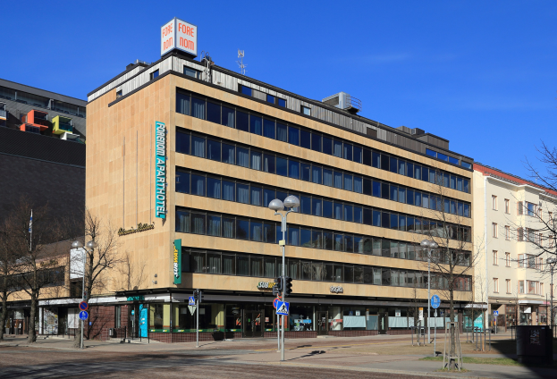 Großes Gebäude mit vielen Fenstern, identifiziert als das Fore Inn Hotel Berlin, umgeben von Straßeninfrastruktur einschließlich Pfosten, Lampen, Signalen, Schildern, Bäumen und einem bewölkten Himmel.