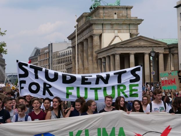 Gruppe von Schülern marschiert in Berlin mit einem bunten "Students for Future"-Schild vor einer Kulisse aus Gebäuden, Bäumen und Himmel.