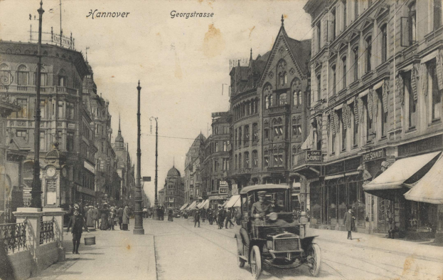 Ein altes Schwarz-Wei√ü-Foto einer Stra√üe in Hannover, Deutschland, mit Fahrzeugen auf der Stra√üe, F√ľ√ügängern auf den Gehwegen, Geb√§uden mit Fenstern auf beiden Seiten, Masten, einer Uhr und einem sichtbaren Himmel.