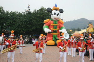 Eine Gruppe von Menschen in Kostümen spielt Musikinstrumente neben einem Mickey-Maus-Floß während eines Umzugs, mit Bäumen, Gebäuden und einem klaren Himmel im Hintergrund; ein Schild mit Text ist links sichtbar.