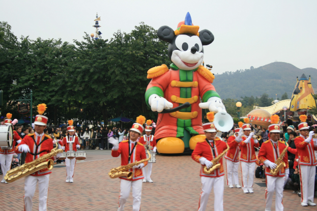 Eine Gruppe von Menschen in Kostümen spielt Musikinstrumente neben einem Mickey-Maus-Floß während eines Umzugs, mit Bäumen, Gebäuden und einem klaren Himmel im Hintergrund; ein Schild mit Text ist links sichtbar.