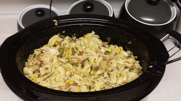 Ein mit Essen gefüllter Slow Cooker steht auf einer Oberfläche mit verschiedenen Tellern und abgeschnittenen Objekten zur Rechten.