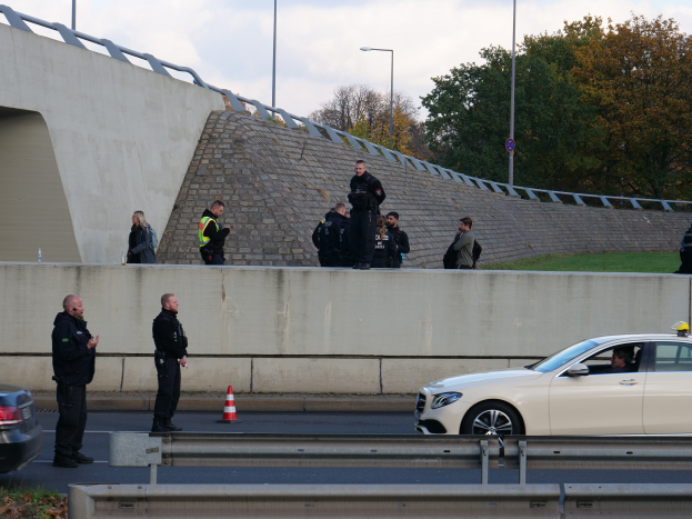 Eine Gruppe von Polizisten steht neben einem Auto auf der Straße, mit Verkehrskegeln, einer Barriere, Gras, einer Wand, Laternenpfählen, Bäumen und einem bewölkten Himmel im Hintergrund.