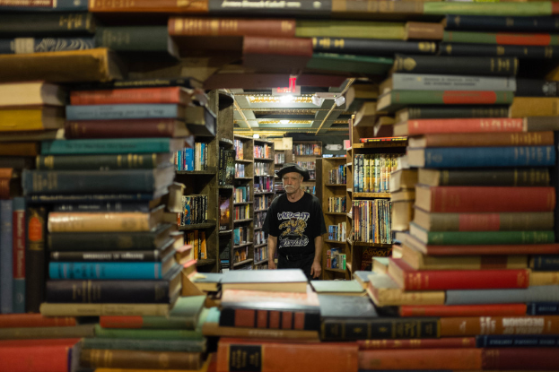 Ein Mann in einer Mütze, T-Shirt und Hose steht vor einem Bücherregal, das ordentlich mit Büchern gefüllt ist.