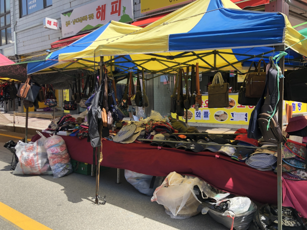 Ein Straßenmarkt mit Zelten, die Taschen, Hüte und andere Artikel präsentieren, vor Häusern mit Fenstern und Texttafeln.