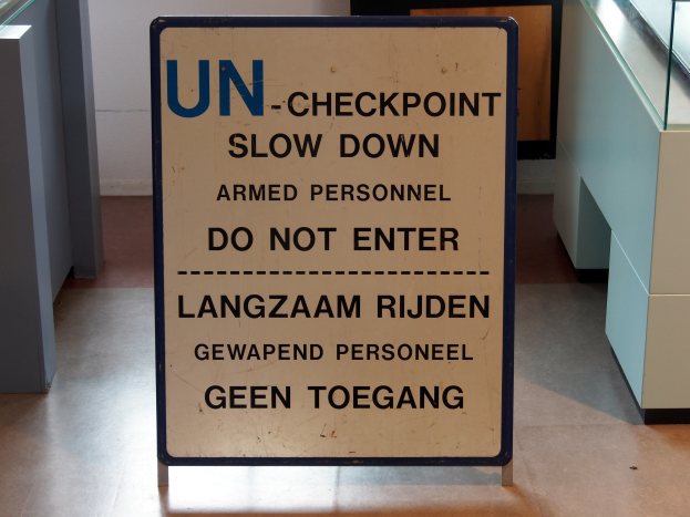 Ein Schild auf dem Boden trägt die Aufschrift "UN-Checkpoint Slow Down Armed Personnel - Do Not Enter" mit einem Glastisch mit verschiedenen Gegenständen darauf zur Rechten und einer Wand im Hintergrund.