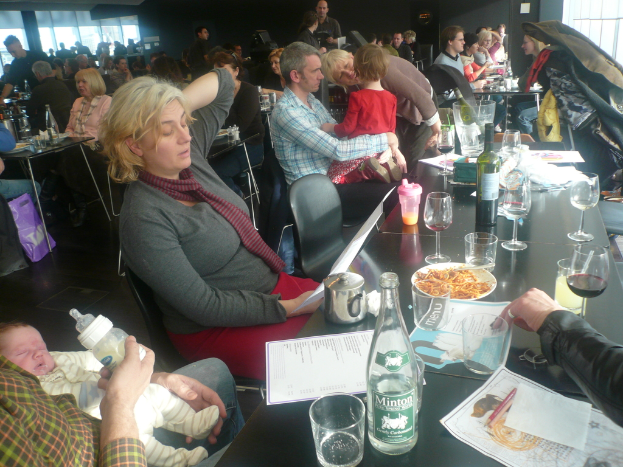 Ein Innenblick auf ein belebtes Restaurant mit Menschen an Tischen, die mit Gläsern, Tellern mit Essen, Getränkeflaschen, Speisekarten und anderen Gegenständen gedeckt sind.