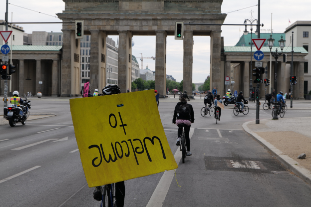 Eine Gruppe von Radfahrern mit Helmen fährt eine Straße entlang vor dem Brandenburger Tor in Berlin, Deutschland, wobei einer ein gelbes Schild hält, Lichtmasten, Verkehrsampeln, Gebäude, Bäume und einen klaren blauen Himmel im Hintergrund.