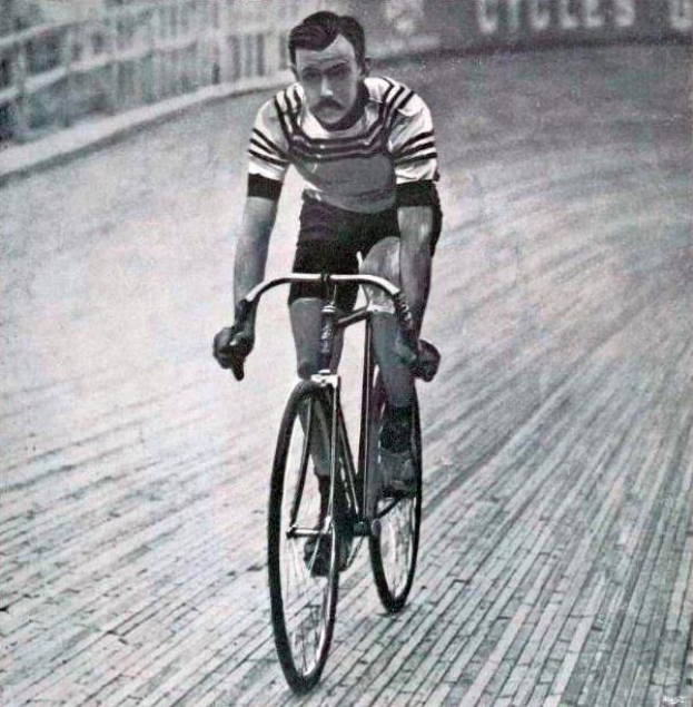 Schwarz-weißes Foto eines Mannes, der auf einer Bahn mit einem Zaun und einer Tafel im Hintergrund Fahrrad fährt.