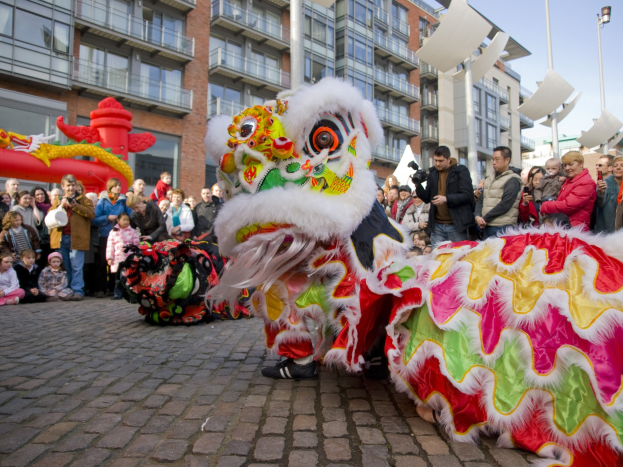 Ein farbenfrohes chinesisches Neujahrsfest in Amsterdam mit einem Löwen Tanz vor einer Zuschauermenge, einige halten Kameras, vor dem Hintergrund von Gebäuden, Laternenmasten und einem klaren blauen Himmel.
