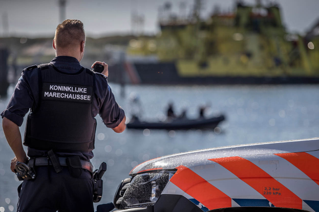 Ein Polizist in Uniform steht neben einem Polizeiwagen in der Nähe eines Gewässers und hält ein Funkgerät in der Hand. Im Hintergrund ist ein Boot mit ein paar Menschen auf dem Wasser zu sehen, unter einem klaren Himmel.