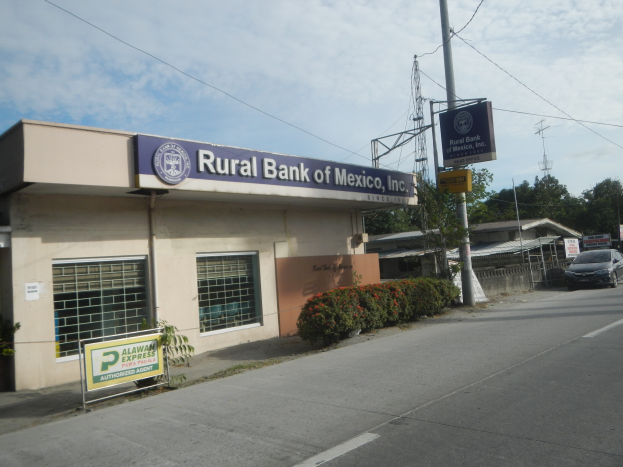 Ein ländliches Gebäude der Bank of Mexico, Inc. mit Fahrzeugen auf der Straße davor, umgeben von Pflanzen mit Blumen, Brettern, Pfählen, Drähten und Häusern, mit Bäumen und einem bewölkten Himmel im Hintergrund.
