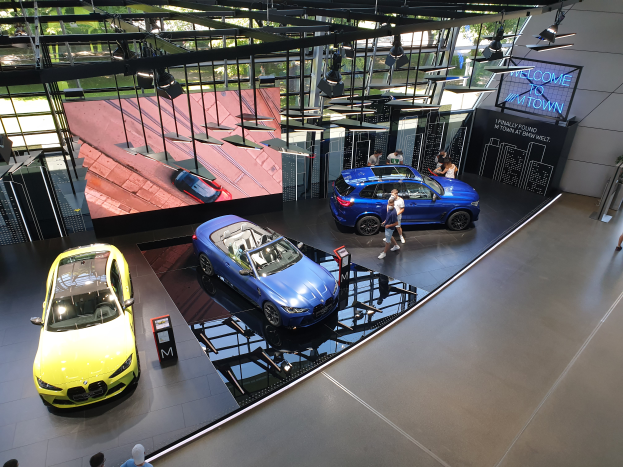 Eine Gruppe von Menschen steht im BMW Museum in München, Deutschland, mit Autos zur Linken und einem Schild mit der Aufschrift "BMW Museum München" zur Rechten, mit Bäumen, Gebäuden und dem Himmel im Hintergrund.
