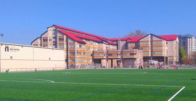 Ein Fußballfeld mit einem großen Gebäude im Hintergrund, das als San Visitor Academy beschriftet ist, umgeben von Bäumen und Laternen, einige Menschen auf dem Feld und der Himmel darüber sichtbar.