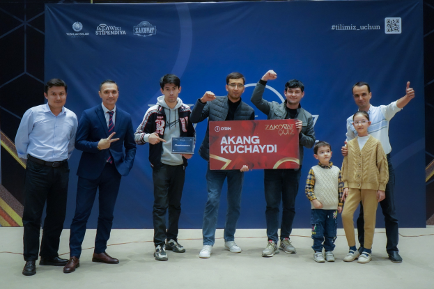 Gruppe von Menschen auf einer Bühne, die eine Tafel mit der Aufschrift "Akang Kuchaydi - Zakovat Quiz" halten, mit einem Banner, Lichtern und anderen Gegenst├Ąnden im Hintergrund.