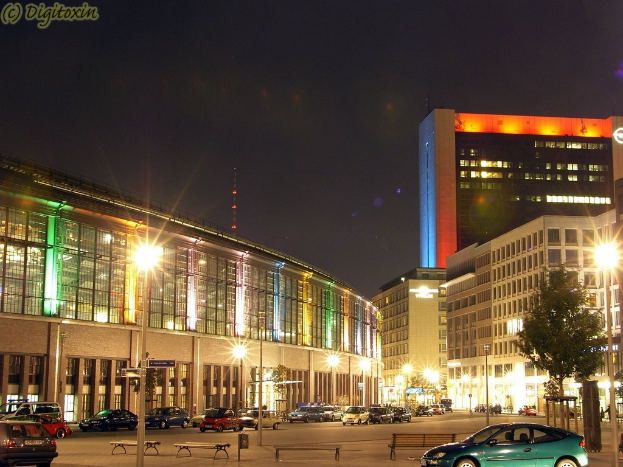 Eine nächtliche Stadtlandschaft mit beleuchteten Gebäuden, Laternen, Bäumen, Bänken und Fahrzeugen auf der Straße, mit Text in der oberen linken Ecke.