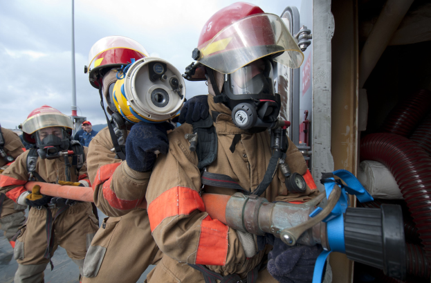 Feuerwehrleute in Schutzausrüstung mit einem Schlauch haltenden, neben einer Stange, Rohren und bewölktem Himmel.