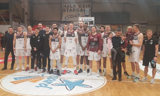 Eine Gruppe von Männern und Kindern auf einem Basketballfeld mit Schildern, die "Hala Osir Starga" lesen, ein Basketballkorb, sitzende Zuschauer und Deckenbeleuchtung.