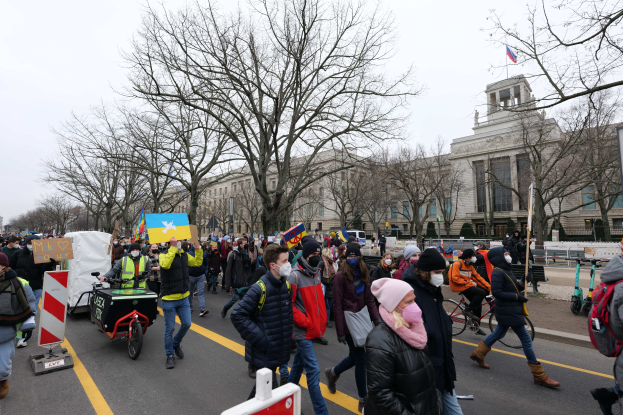 Eine große Gruppe von Menschen marschiert auf einer Straße in Washington, D.C. und hält Schilder und Banner, während einige Fahrräder fahren, unter einem klaren blauen Himmel.