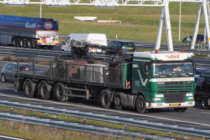 Ein großer Lkw mit Anhänger fährt auf einer Autobahn neben einer Brücke, mit Gras und Geländern auf beiden Seiten, unter einem klaren blauen Himmel mit Masten im Hintergrund.