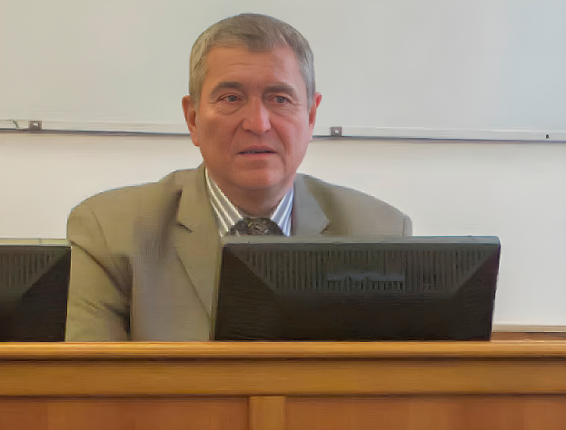 Ein Mann in einem Anzug sitzt an einem Schreibtisch vor einem Computer, mit einer Whiteboard im Hintergrund und einem ernsten Gesichtsausdruck.