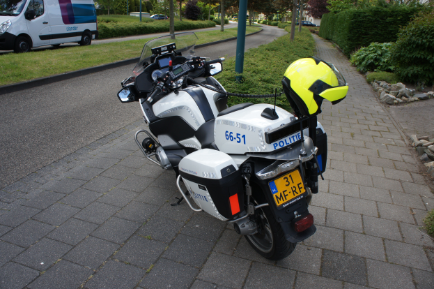 Ein auf der Seite der Straße geparktes Polizeimotorrad mit einem Helm darauf, mit Bäumen, Pfählen, Gebäuden, Pflanzen, Gras, Steinen und anderen Fahrzeugen im Hintergrund.