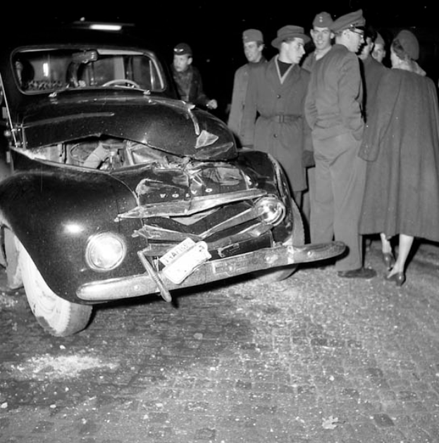 Eine schwarz-weiße Szene eines beschädigten Autos am Straßenrand mit einer Gruppe besorgter Menschen drumherum.