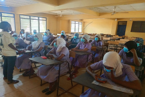 Ein Klassenraum in Nigeria mit Schülerinnen und Schülern, die Gesichtsmasken tragen und auf Bänken sitzen oder stehen, mit Büchern und Gegenständen auf Tischen, Fenstern, einer Tür und Deckenleuchten und -ventilatoren im Hintergrund.