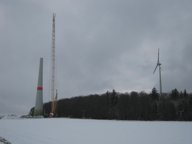 Eine Windkraftanlage steht in einem verschneiten Feld umgeben von Bäumen und Türmen unter einem bewölkten Himmel.