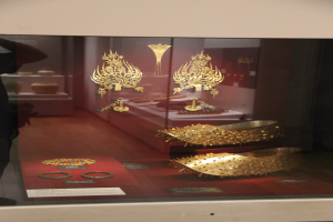 Eine Person in einem schwarzen Kleid steht vor einer Glasvitrine mit glitzerndem Goldschmuck.