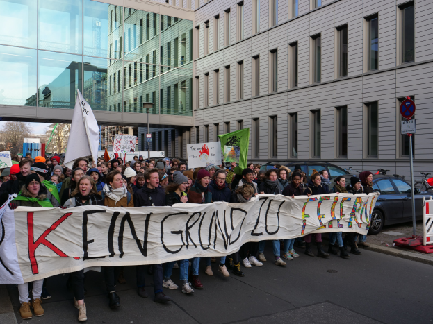 Eine Gruppe von Menschen steht auf der Straße und hält ein Transparent mit der Aufschrift "Kein Grind Lo Feuer" im Protest, mit Gebäuden, Bäumen, Masten, Schildern, Fahrzeugen und einem klaren blauen Himmel im Hintergrund.