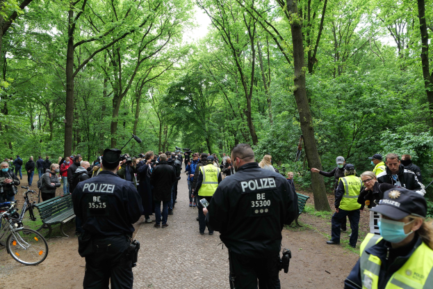 Polizeibeamte in Uniform vor einer Menschenmenge bei einer Outdoor-Veranstaltung mit Fahrrädern und einer Bank im Vordergrund und Bäumen im Hintergrund.