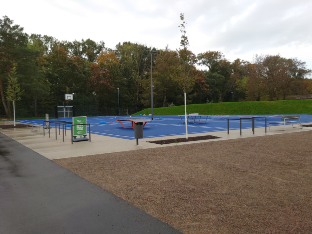Ein Park mit einem Basketballplatz in der Mitte, umgeben von Straßenlaternen, Bänken und einem Mülleimer, mit grünem Gras, Bäumen und einem klaren blauen Himmel.