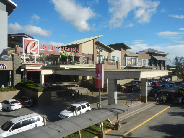 Ein belebter Einkaufszentrum mit verschiedenen Gebäuden, Geschäften, Fahrzeugen auf der Straße, Fußgängern auf dem Gehweg, Straßenlaternen, Schildern, Grünflächen und einem bewölkten Himmel.