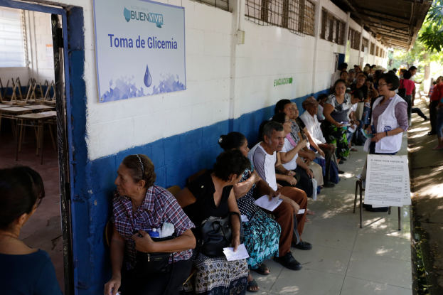 Eine Gruppe von Menschen sitzt auf Bänken vor einem Gebäude mit einer Texttafel an der Wand, einer Tür links und Bäumen im Hintergrund, die auf die Stimmabgabe in einer Wahllokal in Guadalajara, Mexiko, warten