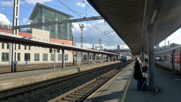 Rechts stehen und sitzen Menschen bei einem Zug, mit Bäumen, Säulen und einem Unterstand; in der Mitte fährt ein Zug auf Schienen mit Texttafeln, einem Unterstand und Säulen; im Hintergrund sind Gebäude, Pfähle und ein bewölkter Himmel zu sehen.