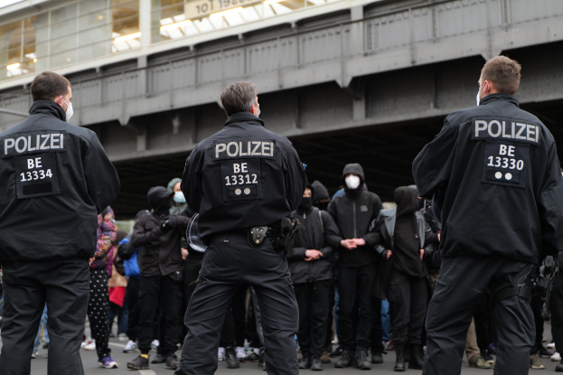Eine Gruppe von Polizisten in Uniform steht vor einer Menge von maskierten Individuen mit einer Brücke und einem Gebäude im Hintergrund bei einer Demonstration in einer Stadt.